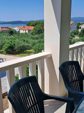 Ein Balkon mit zwei schwarzen Stühlen, einem weißen Geländer und Blick auf Häuser und Grünpflanzen im Hintergrund.