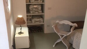 Un dormitorio con una cama, una silla, una lámpara, un estante con cajas de almacenamiento y un espejo pequeño.
