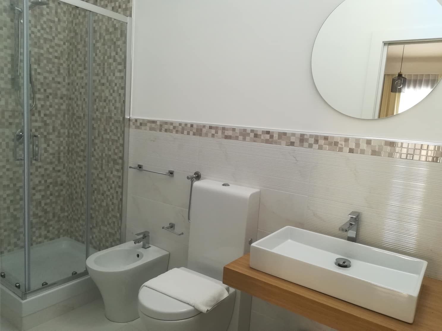 Casa de banho com cabine de chuveiro em vidro, bidé, sanita e lavatório retangular numa bancada de madeira.