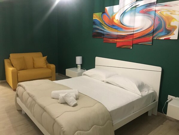 A hotel room with a bed, a yellow armchair, a nightstand with a lamp, and a colorful abstract painting on the wall. - B&B Vittorio Emanuele (Canicatti)