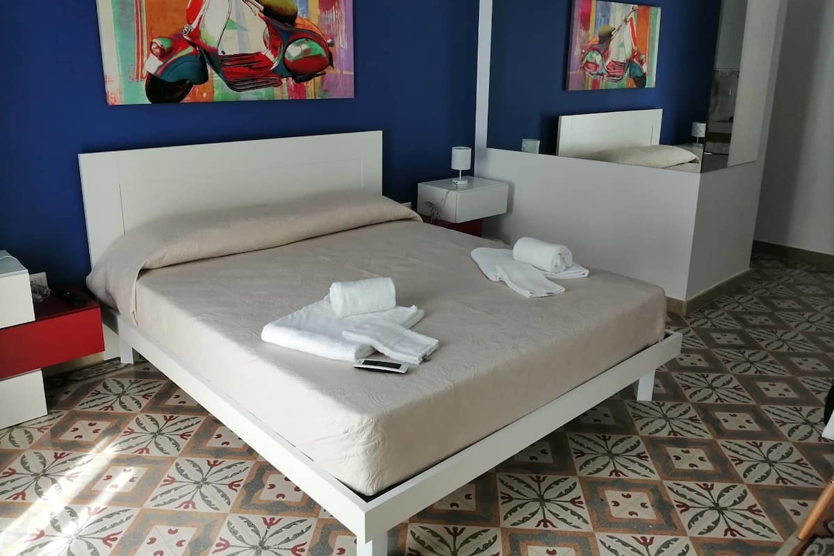 Cama bem arrumada com roupa de cama branca, mesa de cabeceira com abajur e banheiro com espelho e porta-toalhas.
