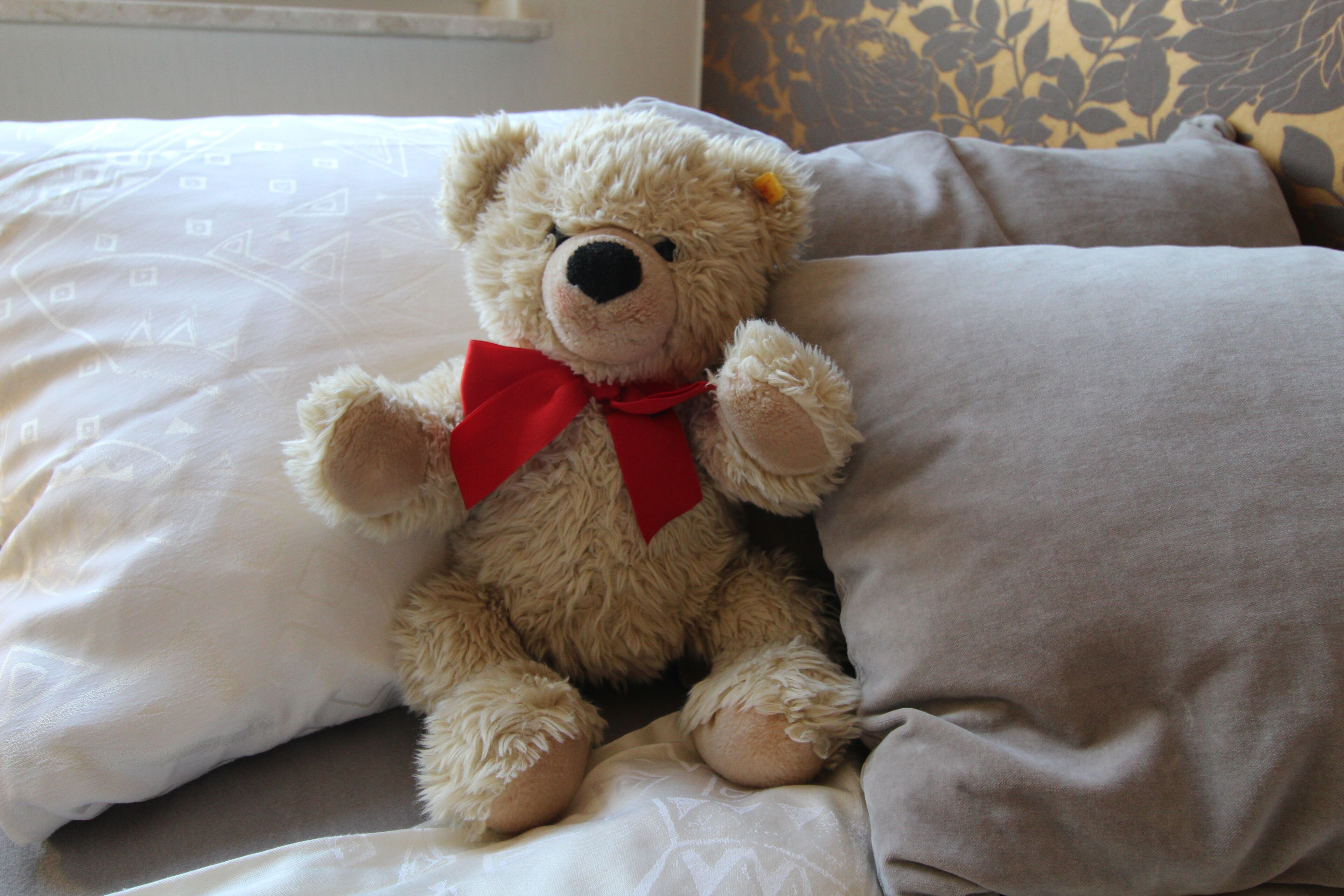 A teddy bear with a red bow sitting on a bed with patterned pillows.