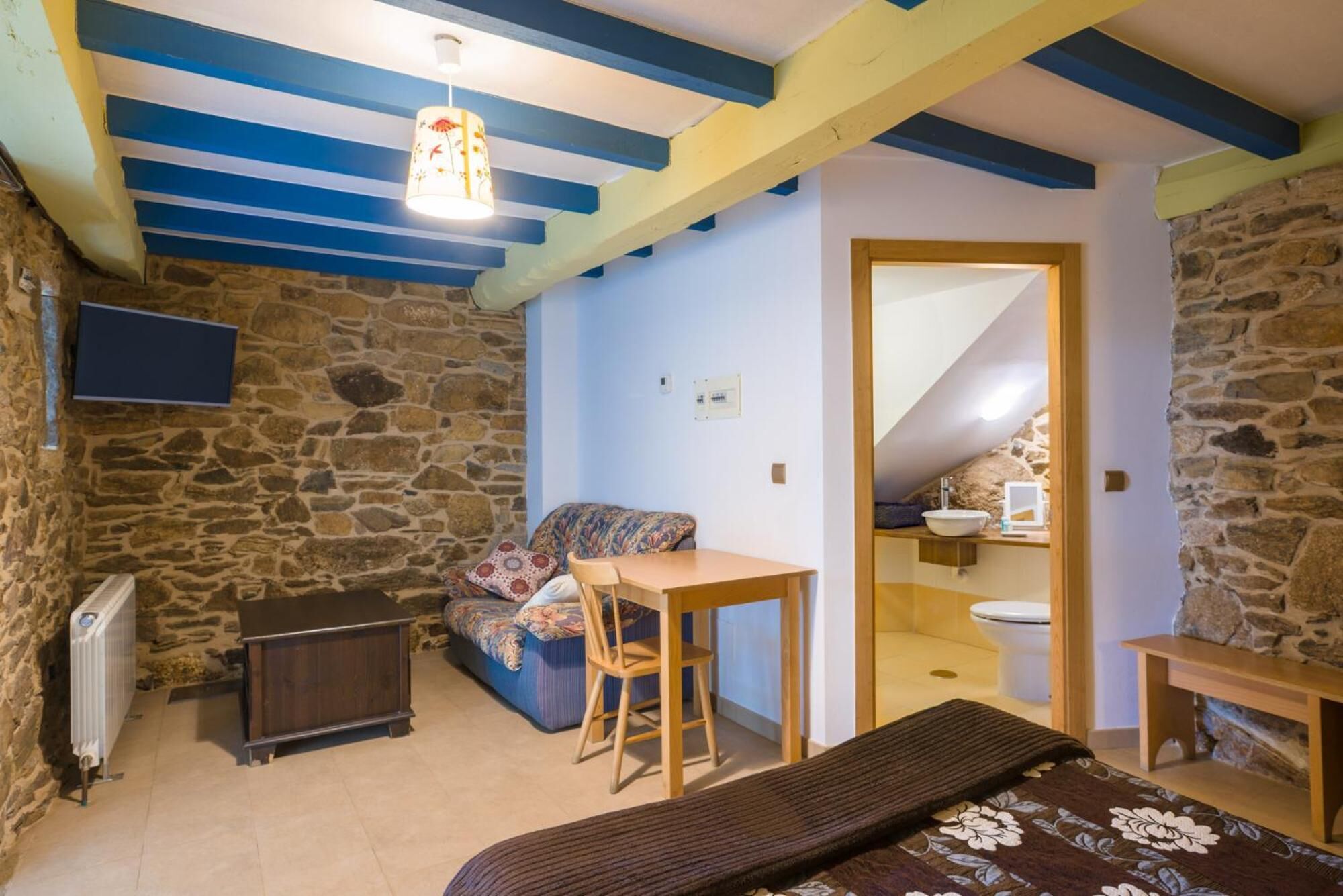 A cozy room with a stone wall, a wooden table, a sofa, and a small bathroom with a sink and toilet.