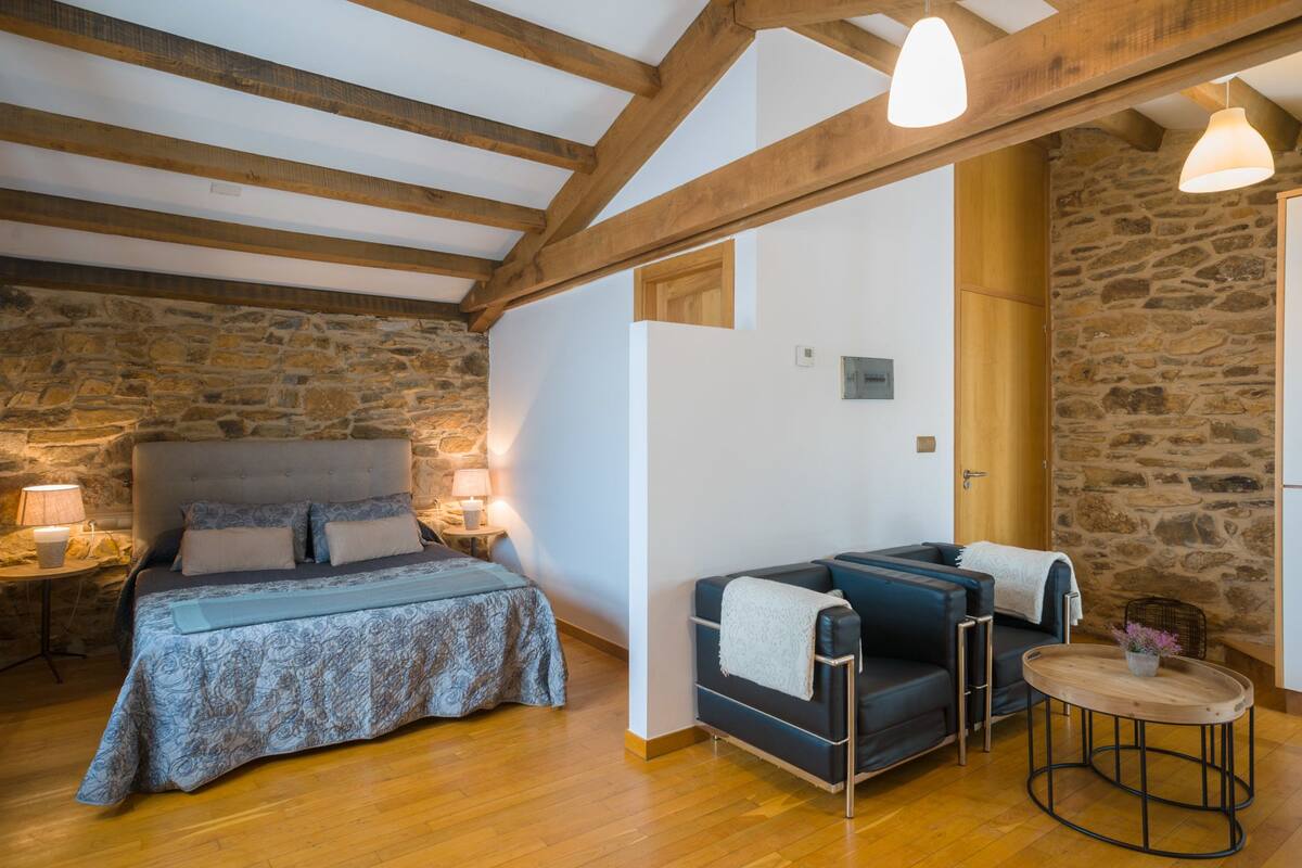 A cozy room with a bed, a sofa, and a small table, featuring exposed wooden beams and stone walls.