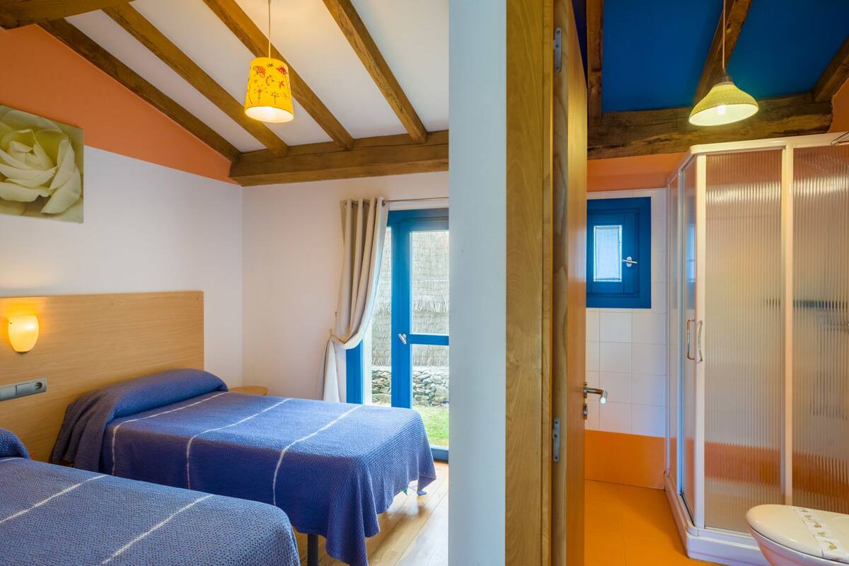 A room with two beds, a wooden ceiling, and a bathroom with a shower and toilet.