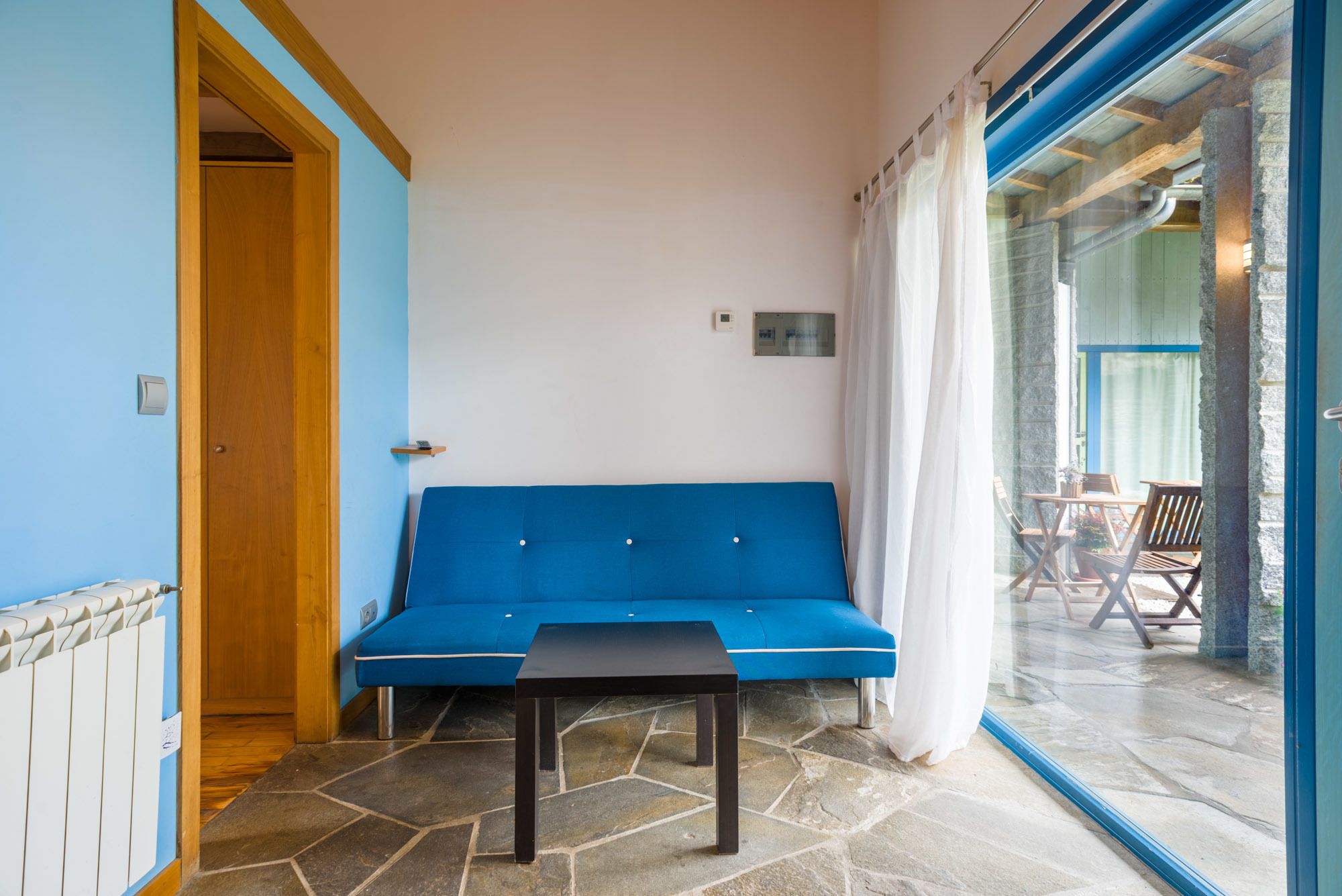Une pièce avec un canapé bleu, une petite table et une porte vitrée donnant sur un espace extérieur équipé d’une table et de chaises.