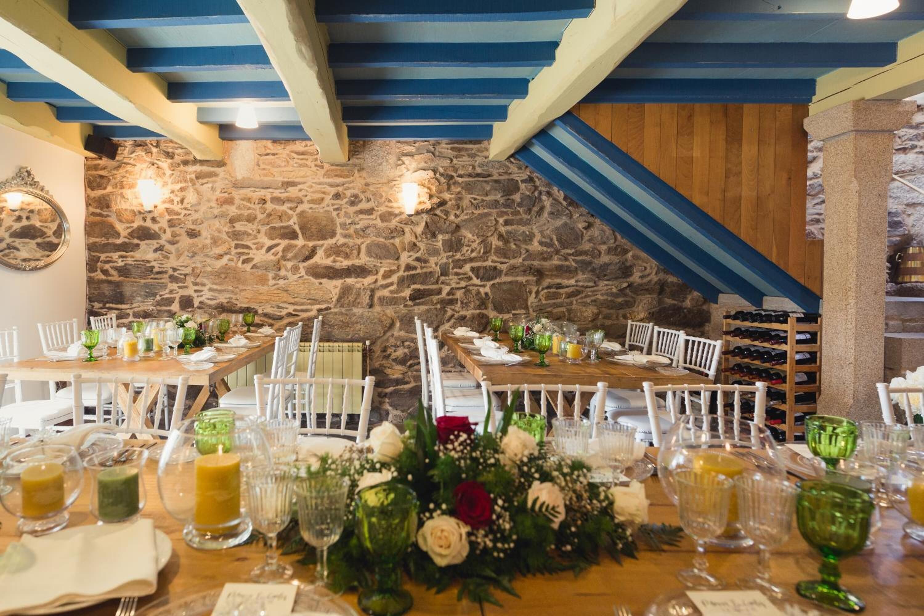 Ein Restaurant mit Steinwänden, Holzbalken und einem Steinfußboden. Die Tische sind mit weißen Stühlen, grünem Glas und floralen Tischdekorationen gedeckt.