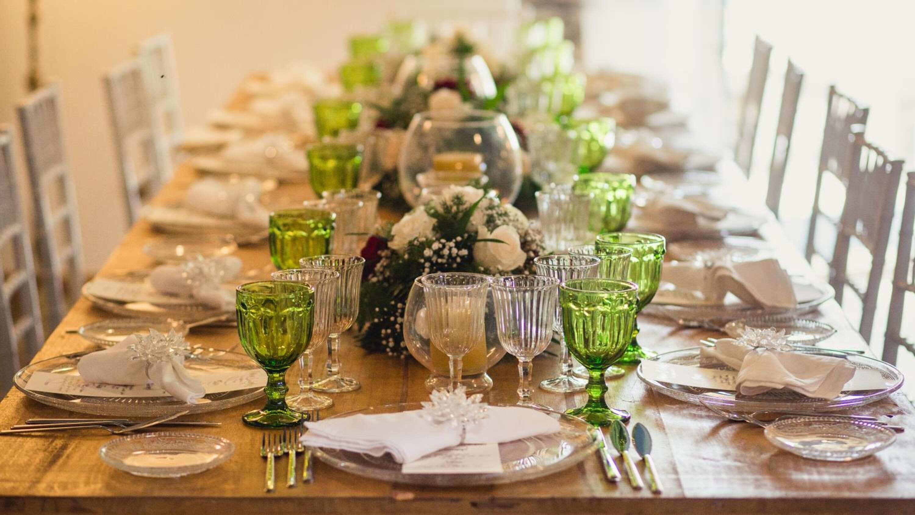 Una larga mesa de comedor puesta con vasos verdes, platos blancos y cubertería.