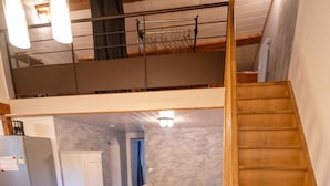 Ampla sala de estar com escada de madeira, sofá e cozinha equipada com geladeira e armários.