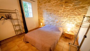 Quarto com parede de pedra, cama com colcha bege, mesa de cabeceira de madeira com abajur e varal de metal.