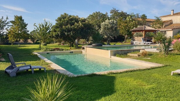 Uma piscina com espreguiçadeiras, rodeada por grama e árvores, e um edifício ao fundo.
