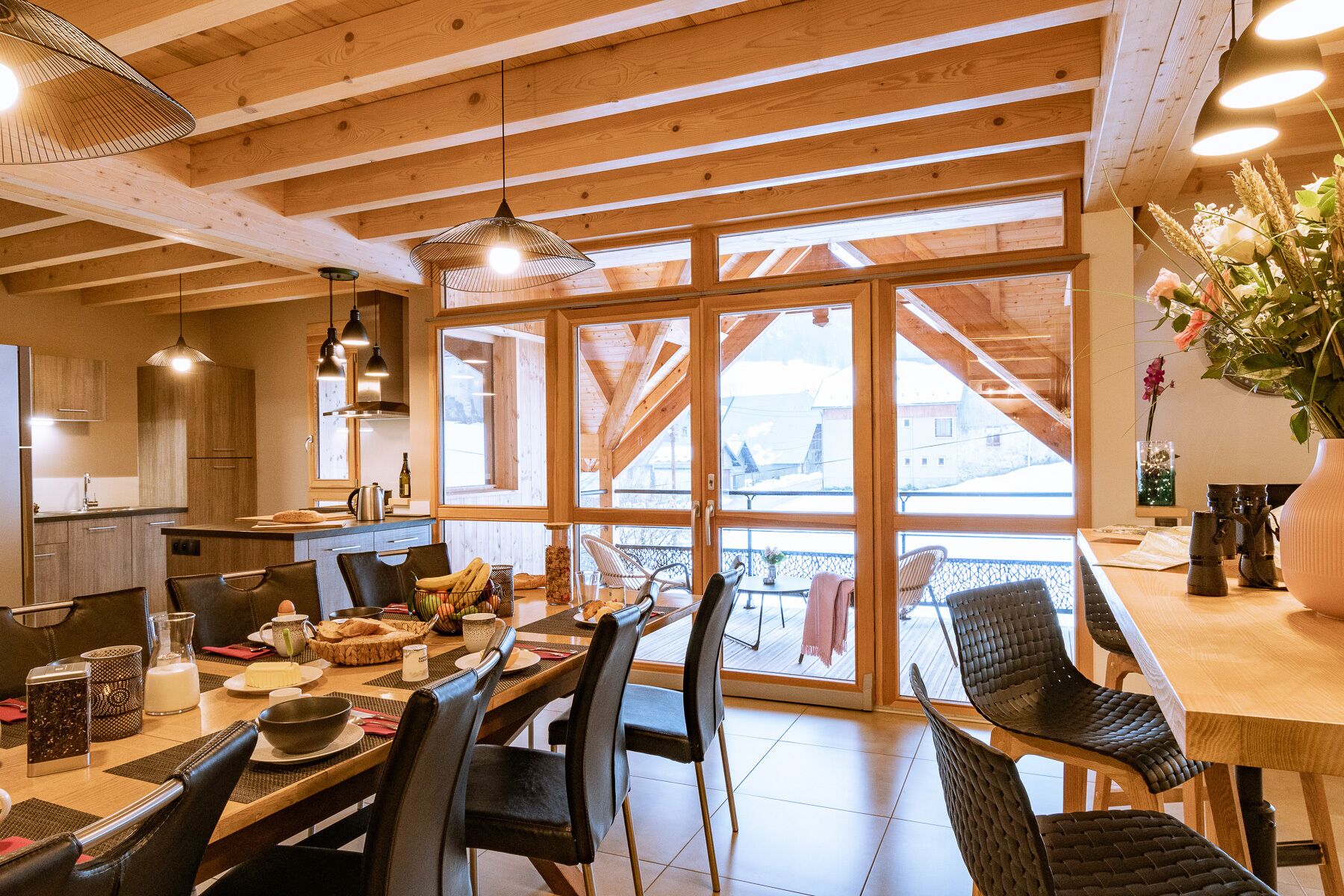 Una cocina moderna con vigas de madera, ventanas amplias y un comedor con sillas y una mesa dispuesta para una comida.