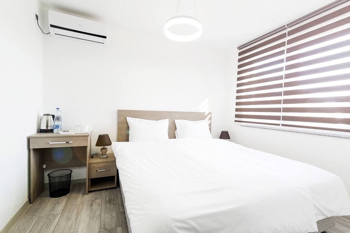 Une chambre d’hôtel comprenant un lit, un bureau avec une chaise, une poubelle, un appareil de climatisation, une lampe et une fenêtre avec des stores.