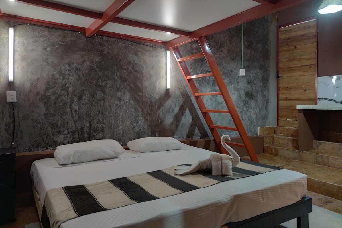 Un dormitorio con cama, almohadas, manta a rayas, escalera de madera y pared con superficie texturizada.
