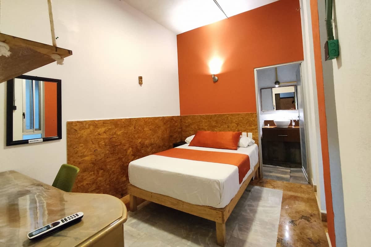 Habitación de hotel con cama, escritorio, silla, espejo y baño visible a través de una puerta abierta.