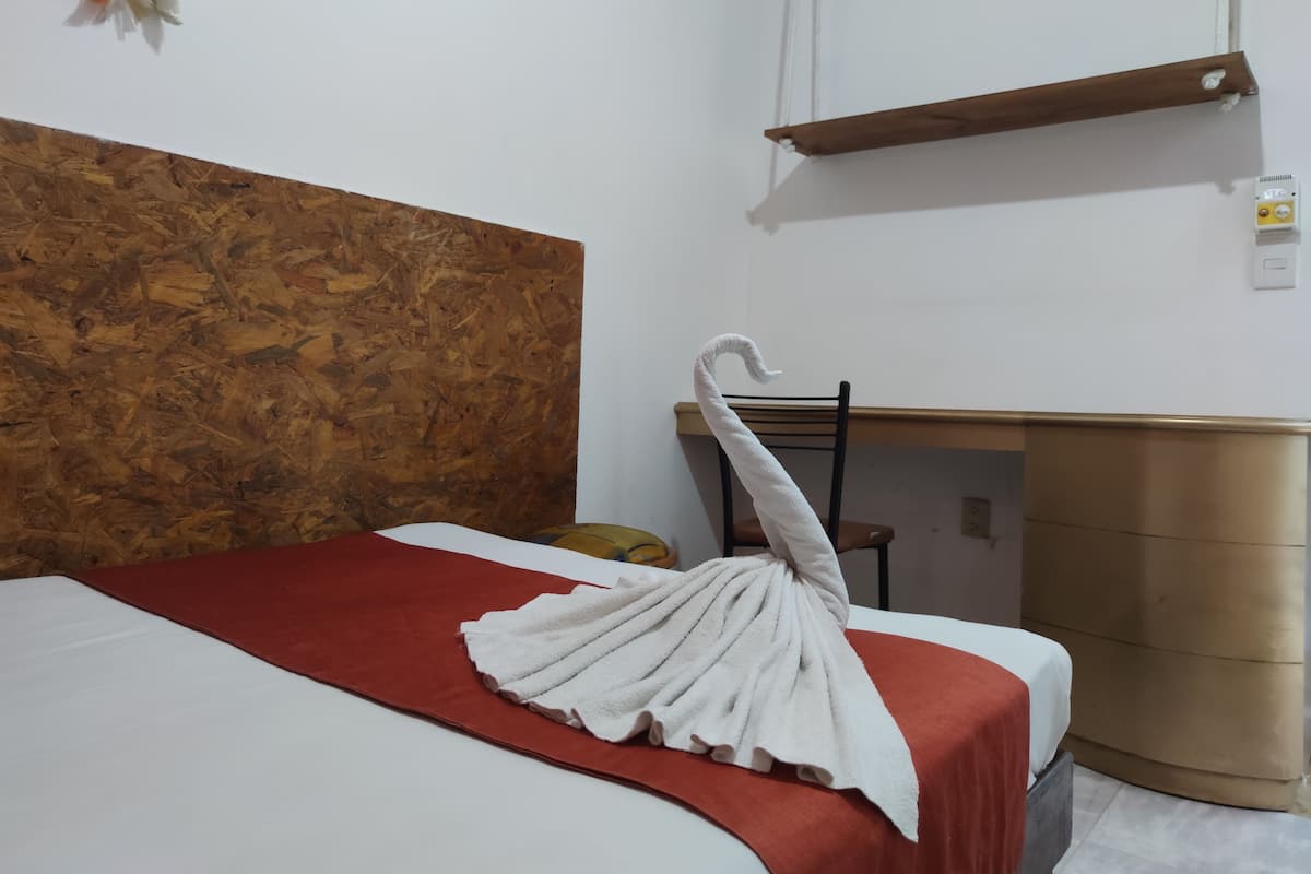Habitación de hotel con cabecera de madera, cama con colcha roja y blanca, toalla blanca sobre la cabecera, una silla de madera y un pequeño estante de madera.