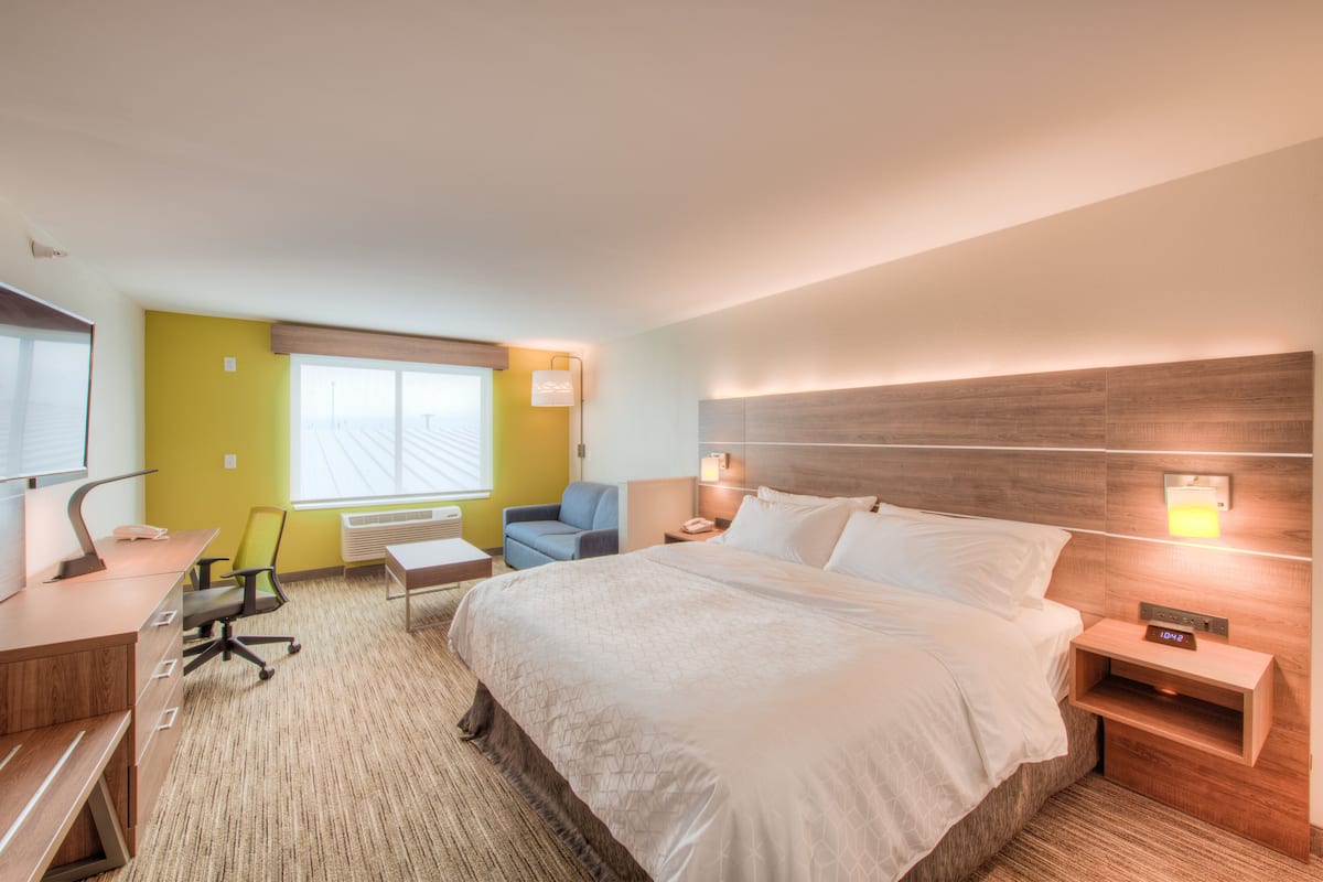 A hotel room with a large bed, a desk with a chair, a small table, and a window with blinds.
