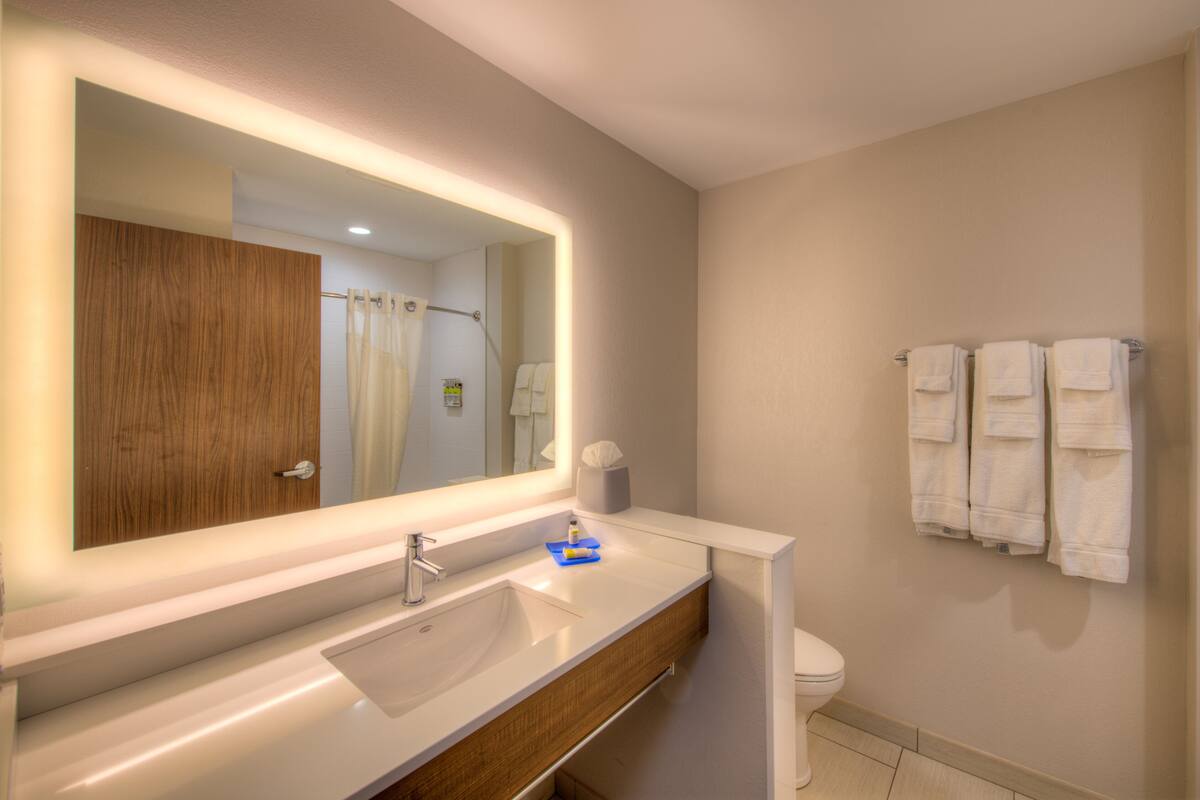 A modern bathroom with a large mirror, white sink, wooden vanity, and a toilet with towels hanging on the wall.