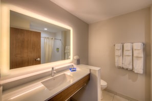 A modern bathroom with a large mirror, white sink, wooden vanity, and a toilet with towels hanging on the wall.