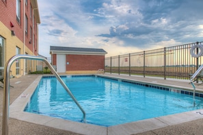 A swimming pool with a metal safety railing, a lifebuoy, and a brick building in the background.