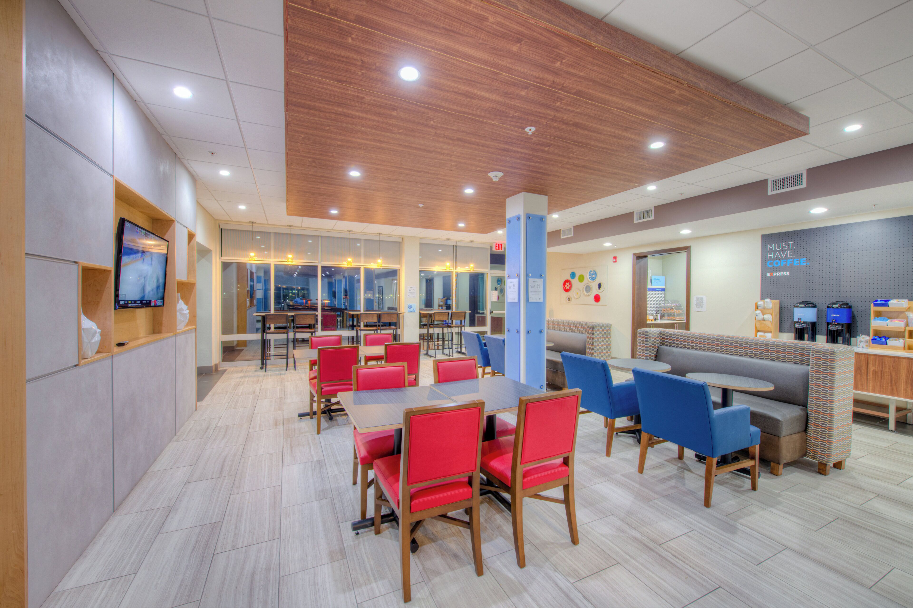 Una cafetería moderna con asientos rojos y azules, una estación de café y un televisor en la pared.
