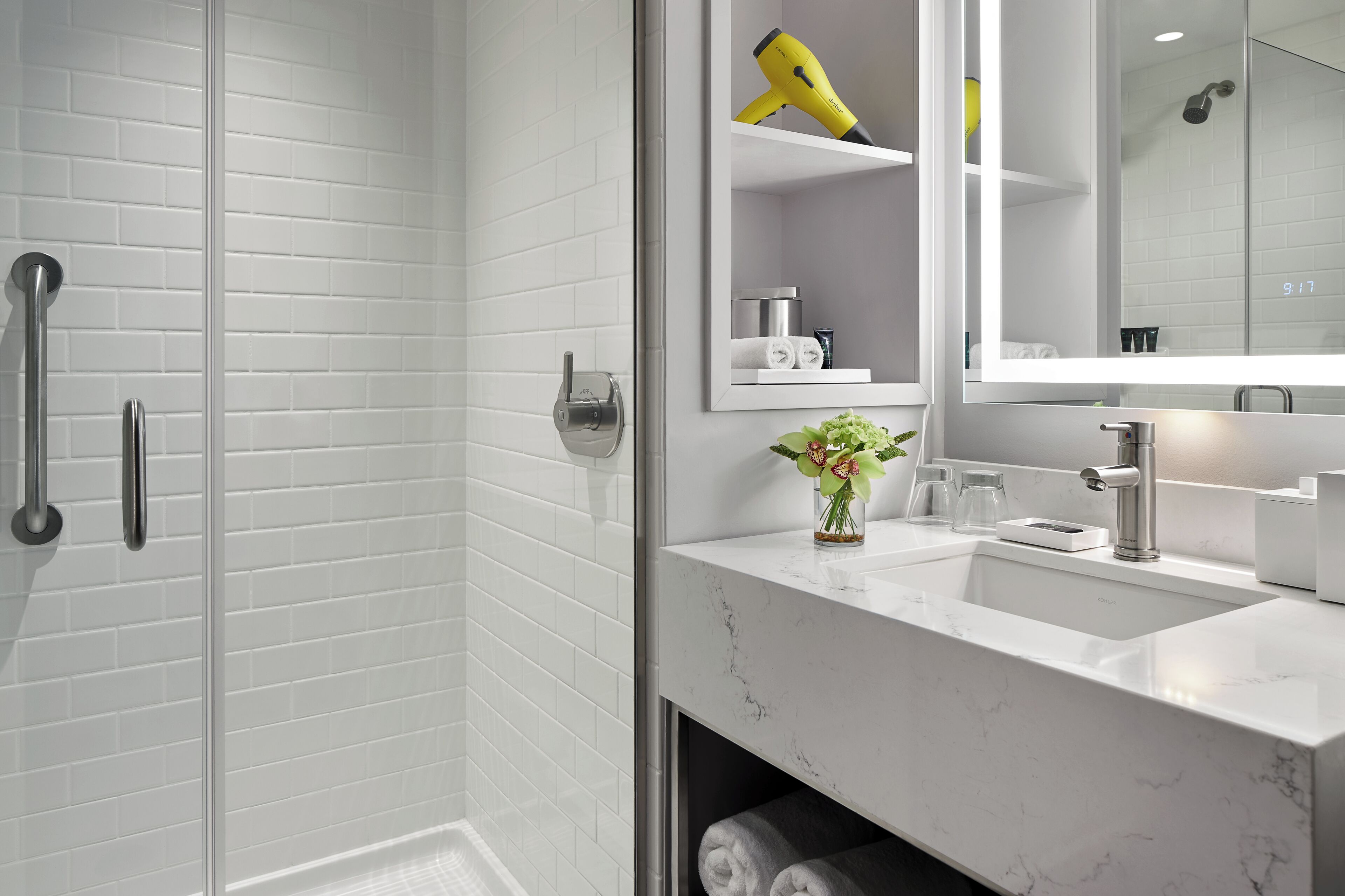 A modern bathroom with a white subway tile wall, a glass shower enclosure, a white sink with a marble countertop, a mirror, and a yellow hairdryer on a shelf.