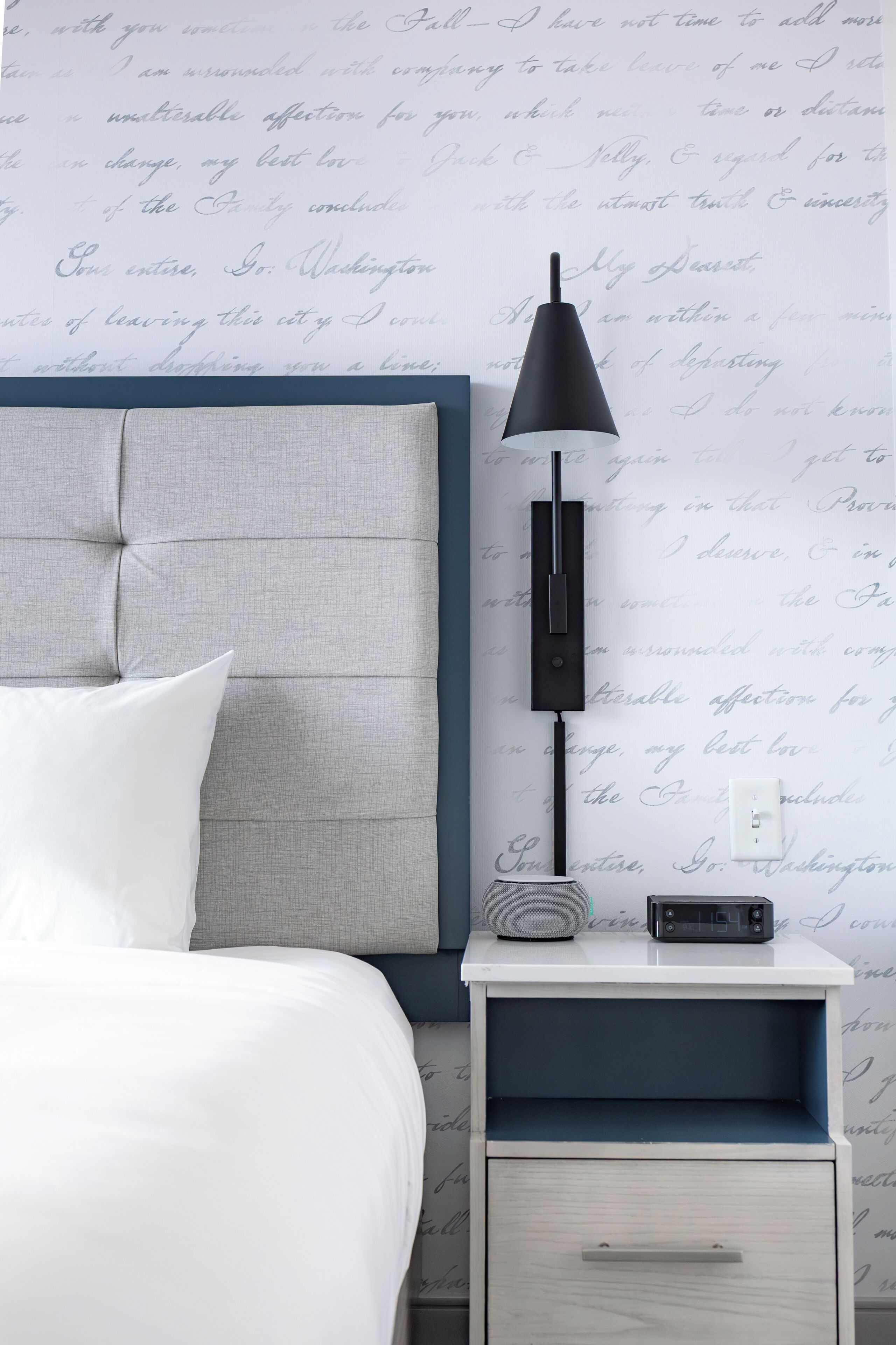 A neatly made bed with white linens, a grey upholstered headboard, a bedside table with a black lamp, and a small speaker.