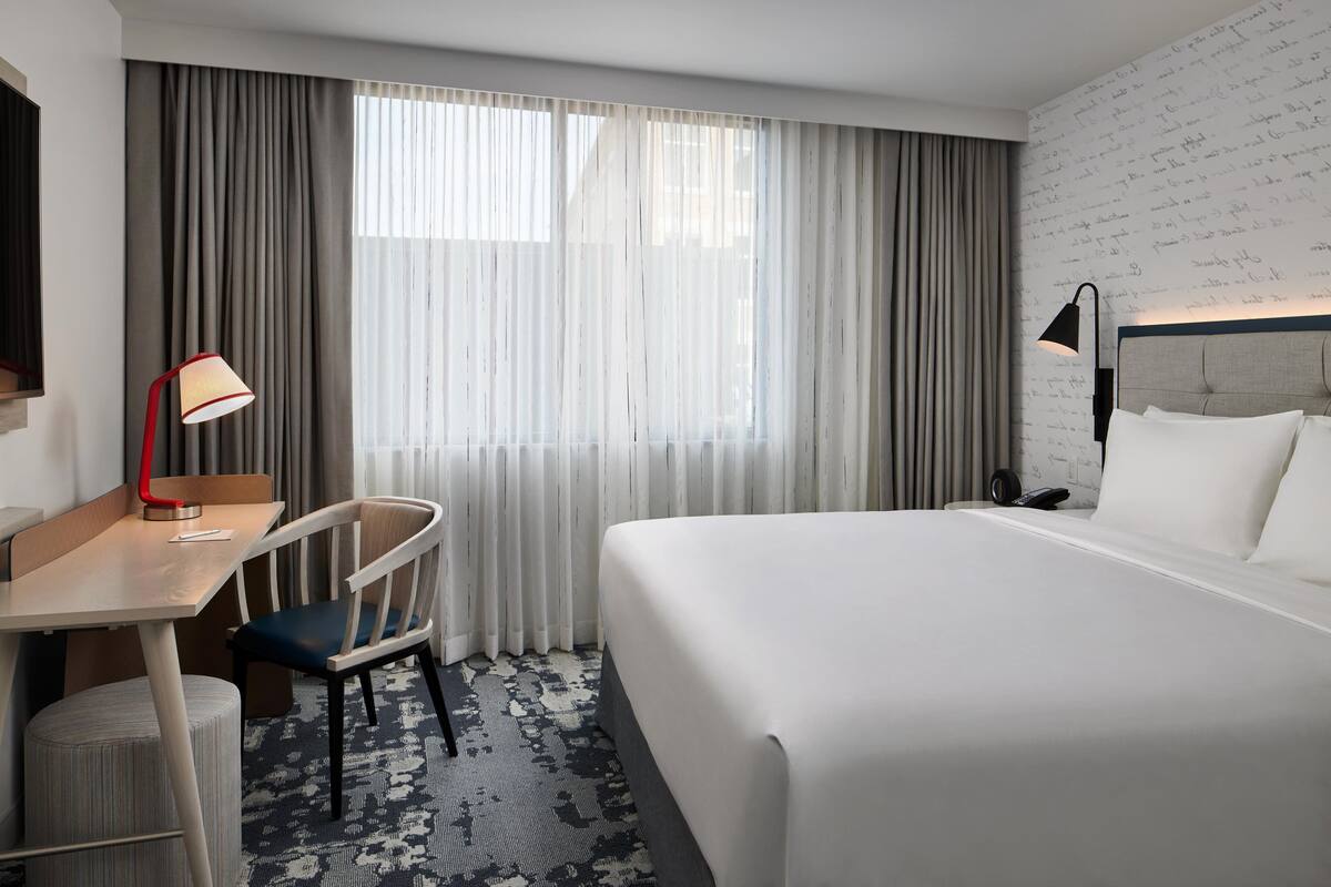 Habitació d'hotel amb un llit gran, un escriptori amb cadira, una làmpada i una finestra amb cortines.