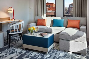 Una sala de estar moderna con un sofá gris, un reposapiés azul y una silla de madera. Hay una mesita con un libro y un jarrón de flores.