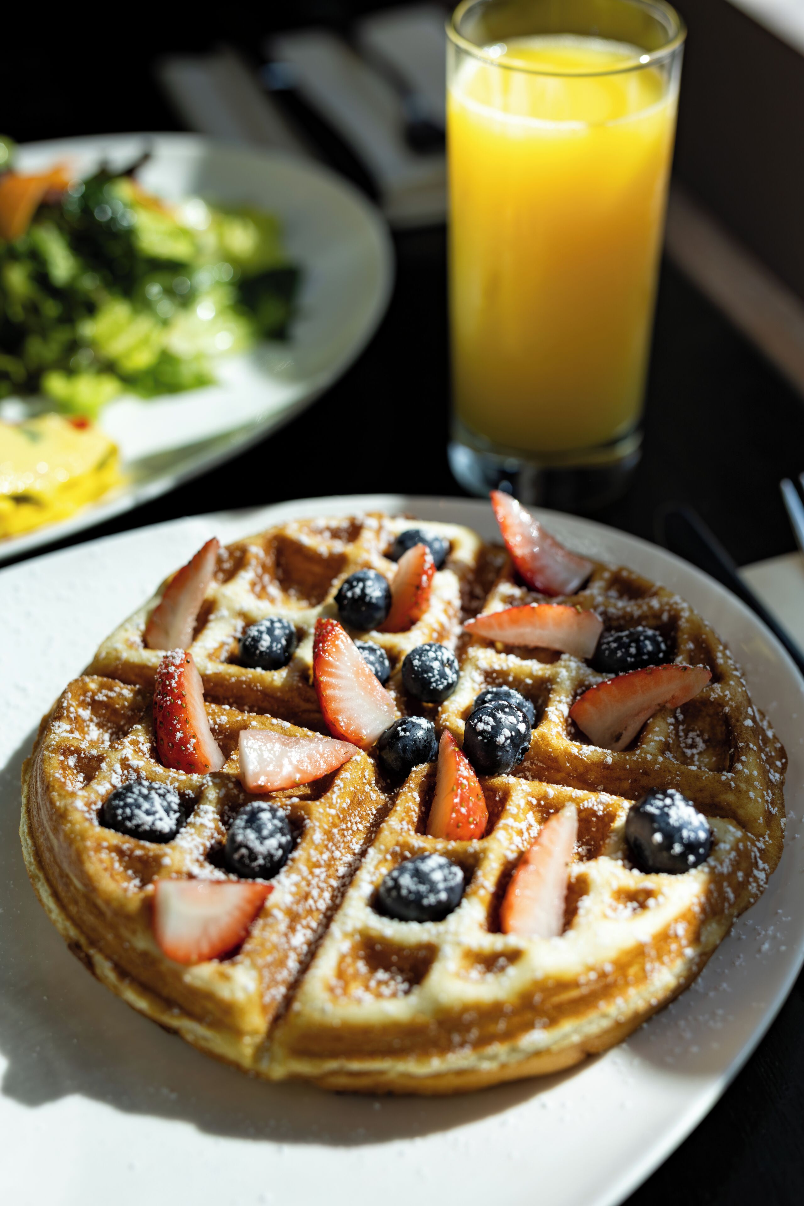 Une assiette de petit-déjeuner avec un gaufre garnie de fraises et de myrtilles, un verre de jus d’orange et une portion de légumes verts.