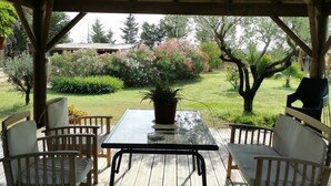 Une terrasse couverte, dotée d’une table et de chaises, entourée d’un jardin où poussent différentes plantes et arbres.