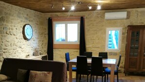 Un salon chaleureux avec un mur en pierre, un plafond en bois, un coin repas avec une table et des chaises, et une fenêtre avec des rideaux.