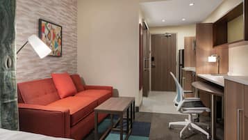 A hotel room with a red sofa, a small table, a desk with a chair, a TV, and a painting on the wall.