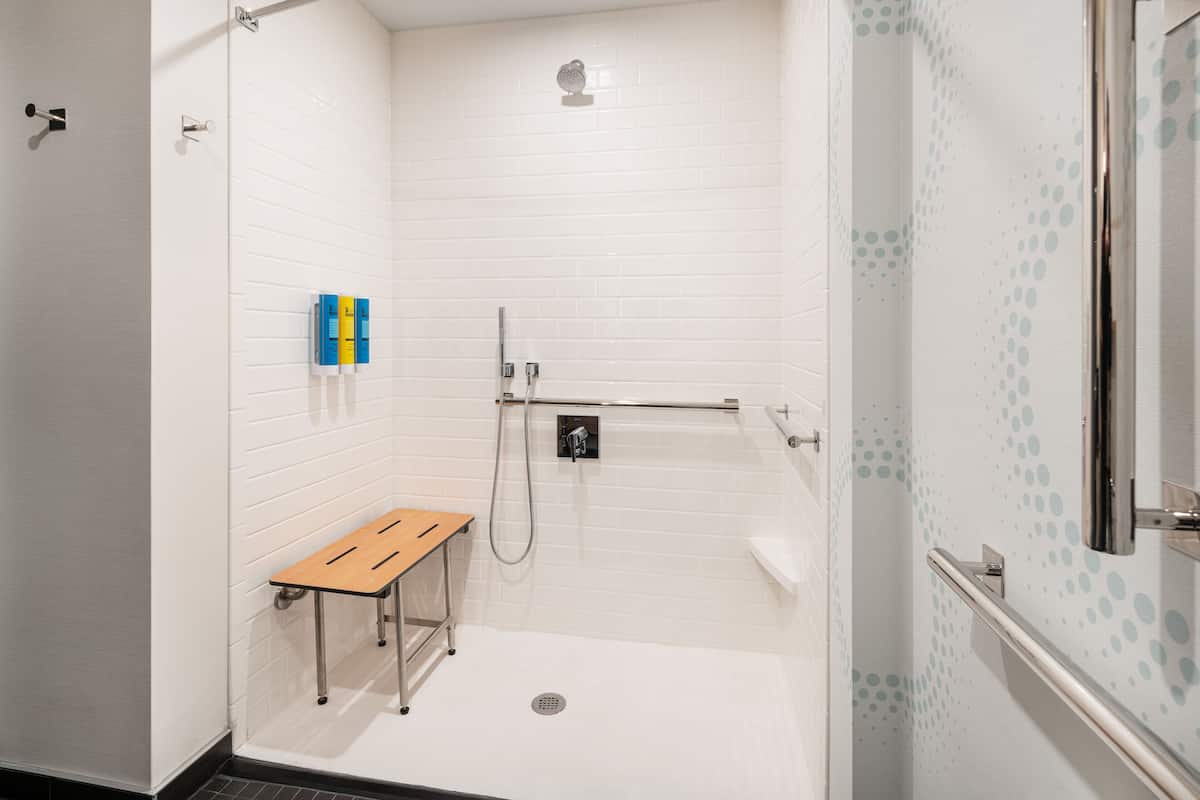 Un baño moderno con paredes de azulejos blancos, ducha con cabezal de ducha de mano, un banco y barra de apoyo.