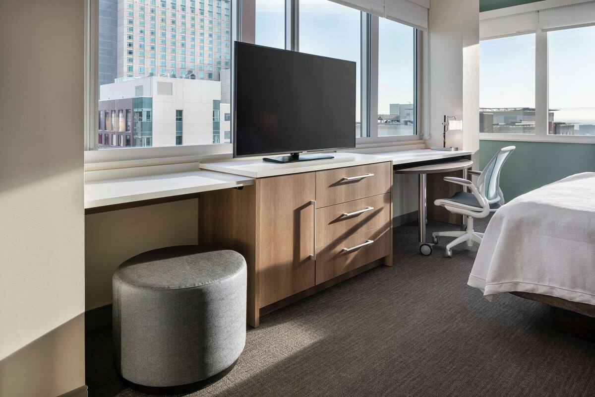 Una habitación de hotel moderna con un ventanal grande, un televisor de pantalla plana, un escritorio de madera, un taburete gris y una cama con cubrecama blanco.