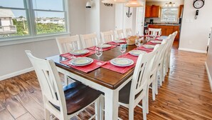 A dining room with a wooden table set for six, white chairs, and a view of the backyard through large windows.