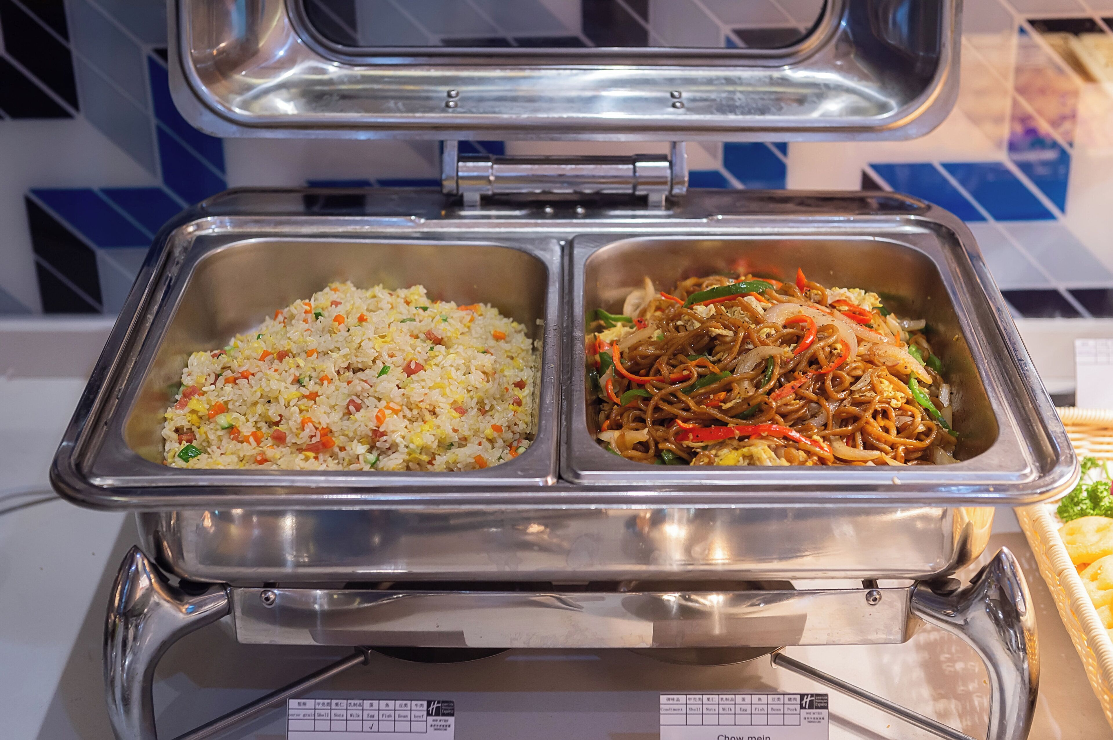 A buffet with two metal serving trays, one containing fried rice and the other noodles with vegetables.