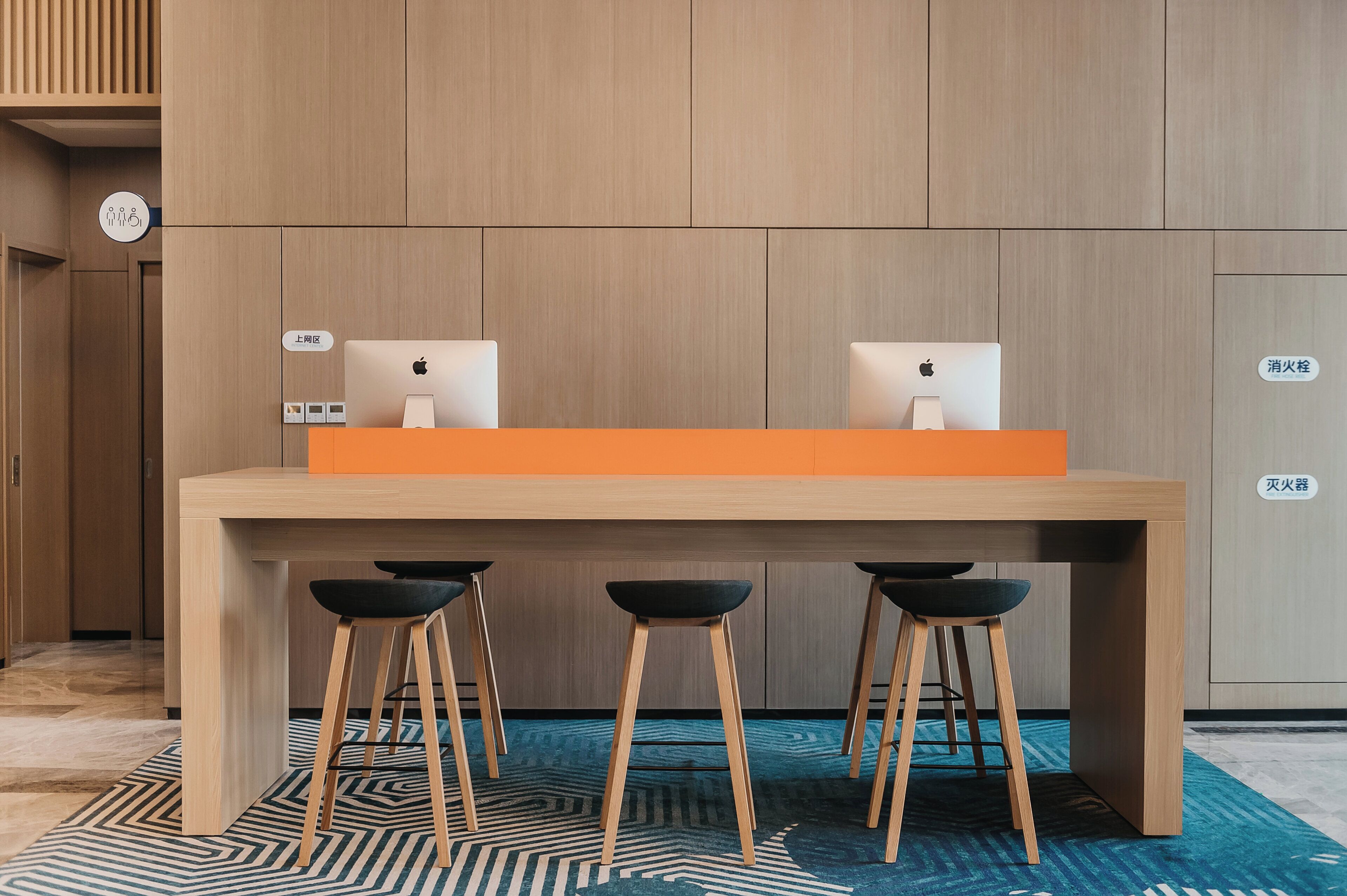 A modern hotel lobby with a wooden reception desk, two Apple iMac computers, and three black bar stools.