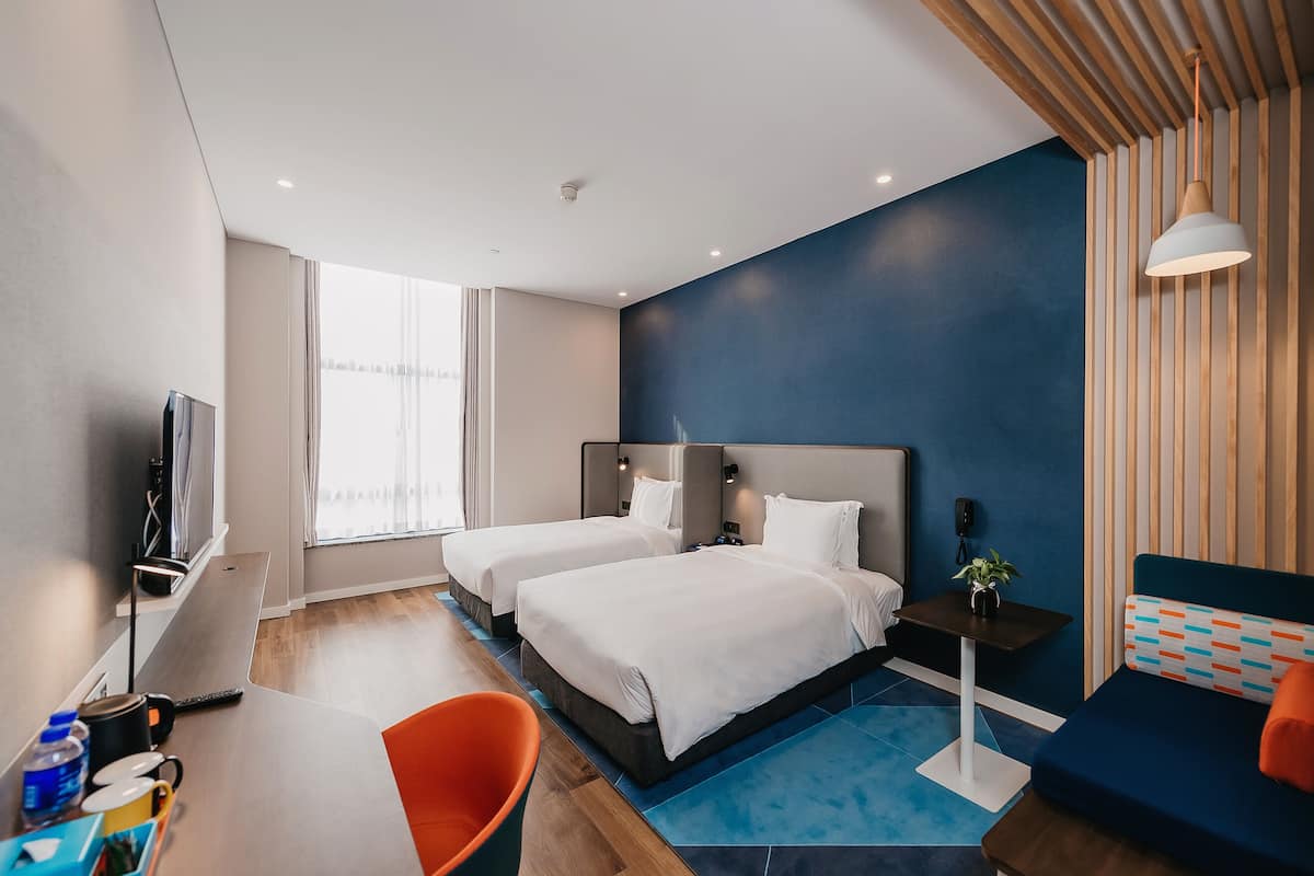 Habitación de hotel con dos camas, un escritorio con silla, televisión y un ventanal grande.