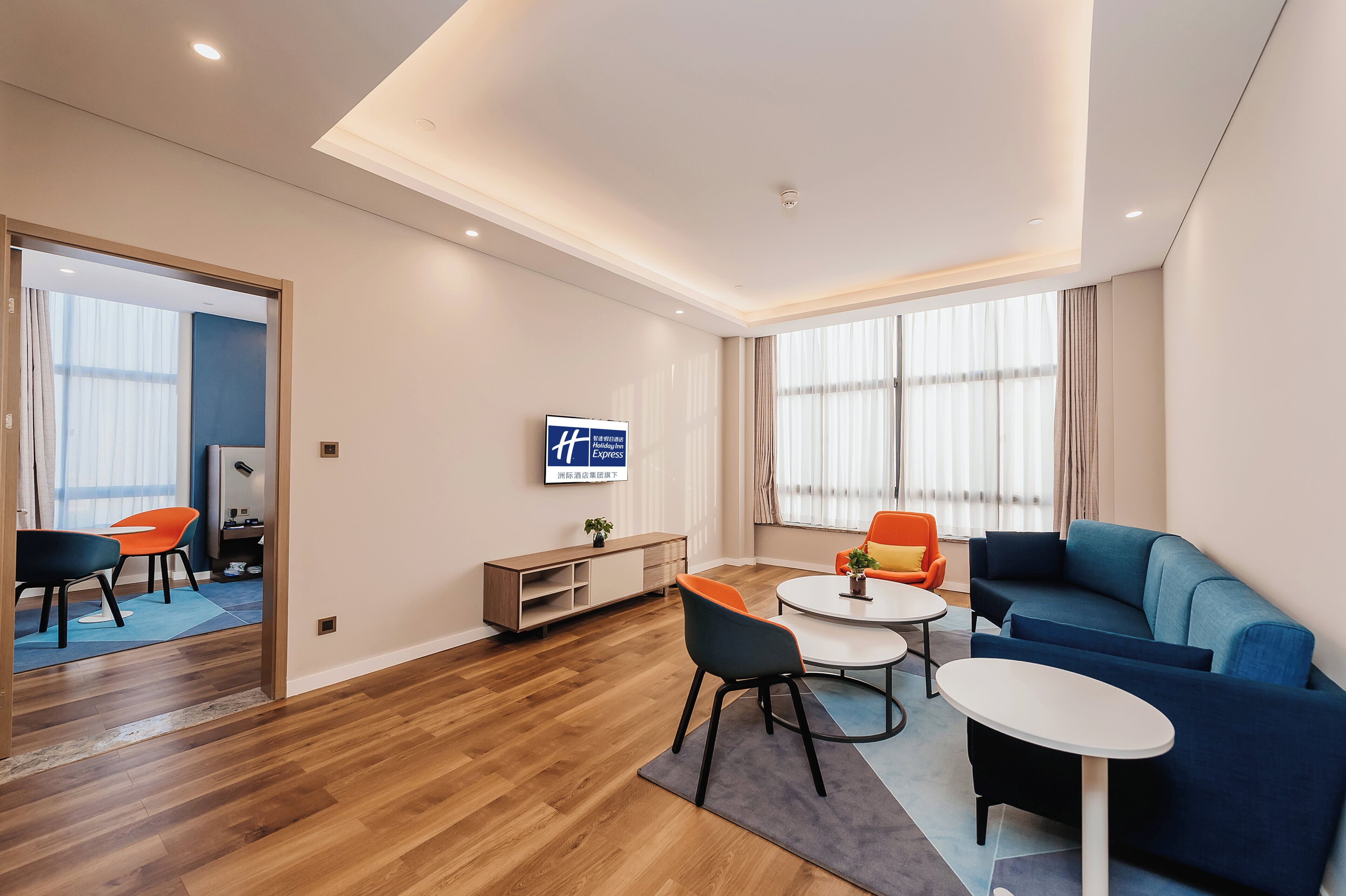 A modern hotel room with a wooden floor, a blue sofa, a round coffee table, and a TV mounted on the wall.