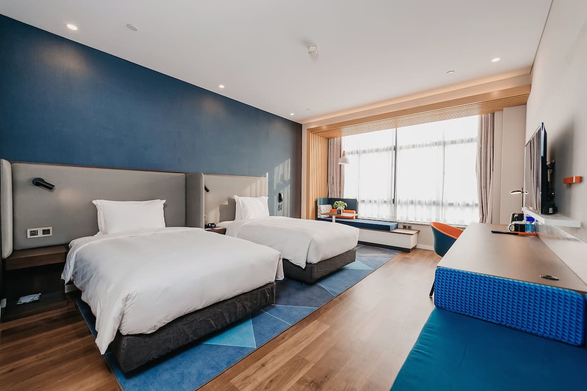 Una habitación de hotel moderna con dos camas, un escritorio, un televisor y un amplio ventanal con cortinas.