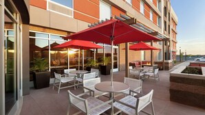 An outdoor patio area with round tables and white wicker chairs, red umbrellas, and potted plants. - Home2 Suites by Hilton Scottsdale Salt River (Scottsdale)
