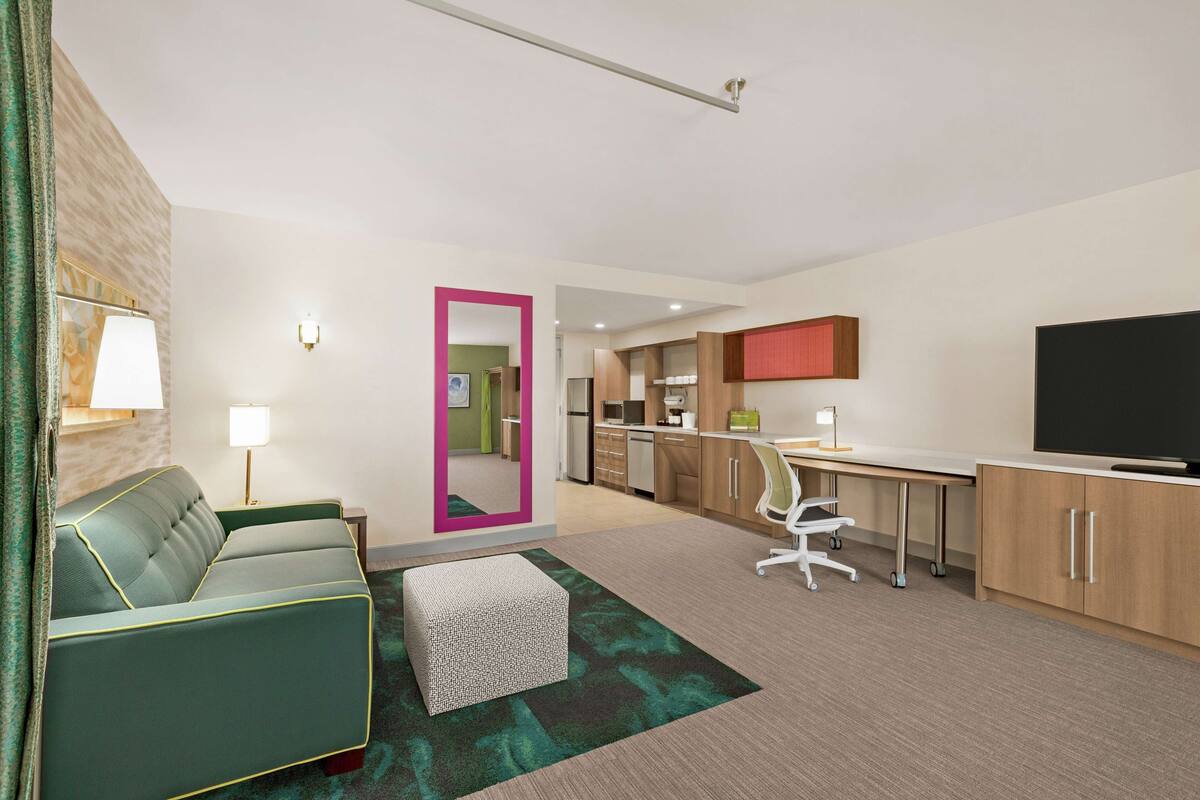 Een moderne hotelkamer met een groene bank, een klein poufje, een bureau met stoel, een flatscreen televisie en een keukenhoek.