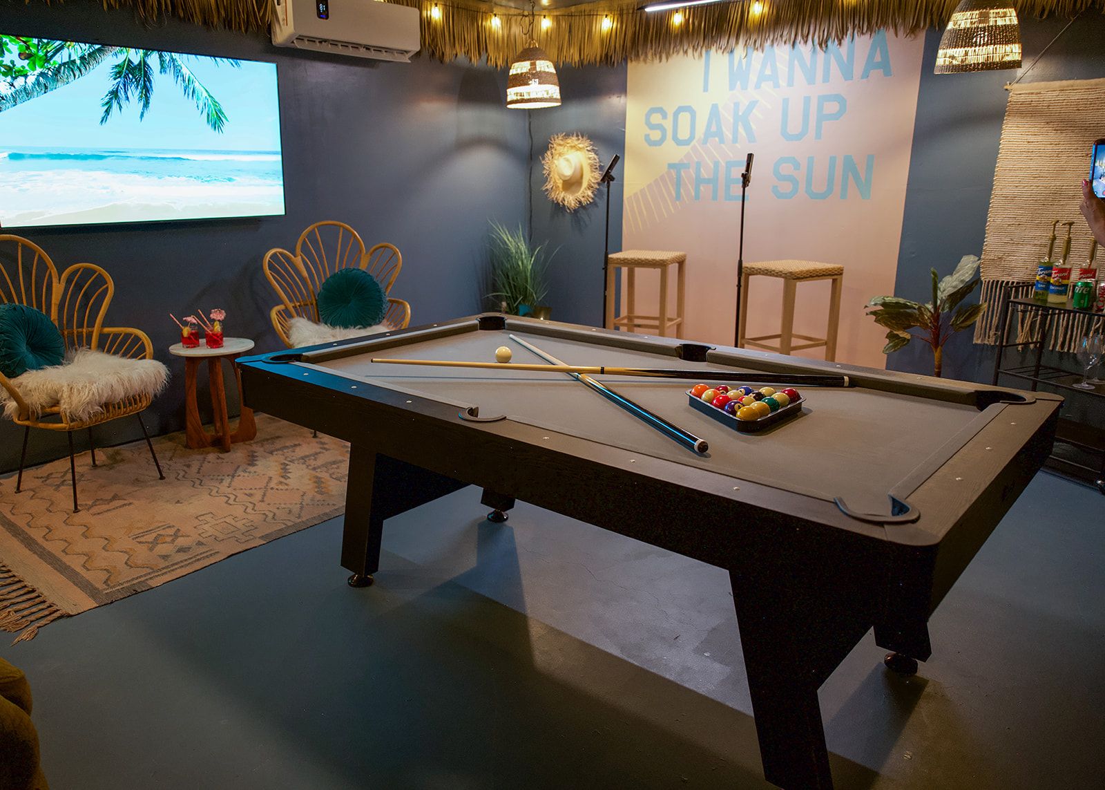 A pool table in a room with a tropical-themed wall mural, a projector screen displaying a beach scene, and a pool cue.
