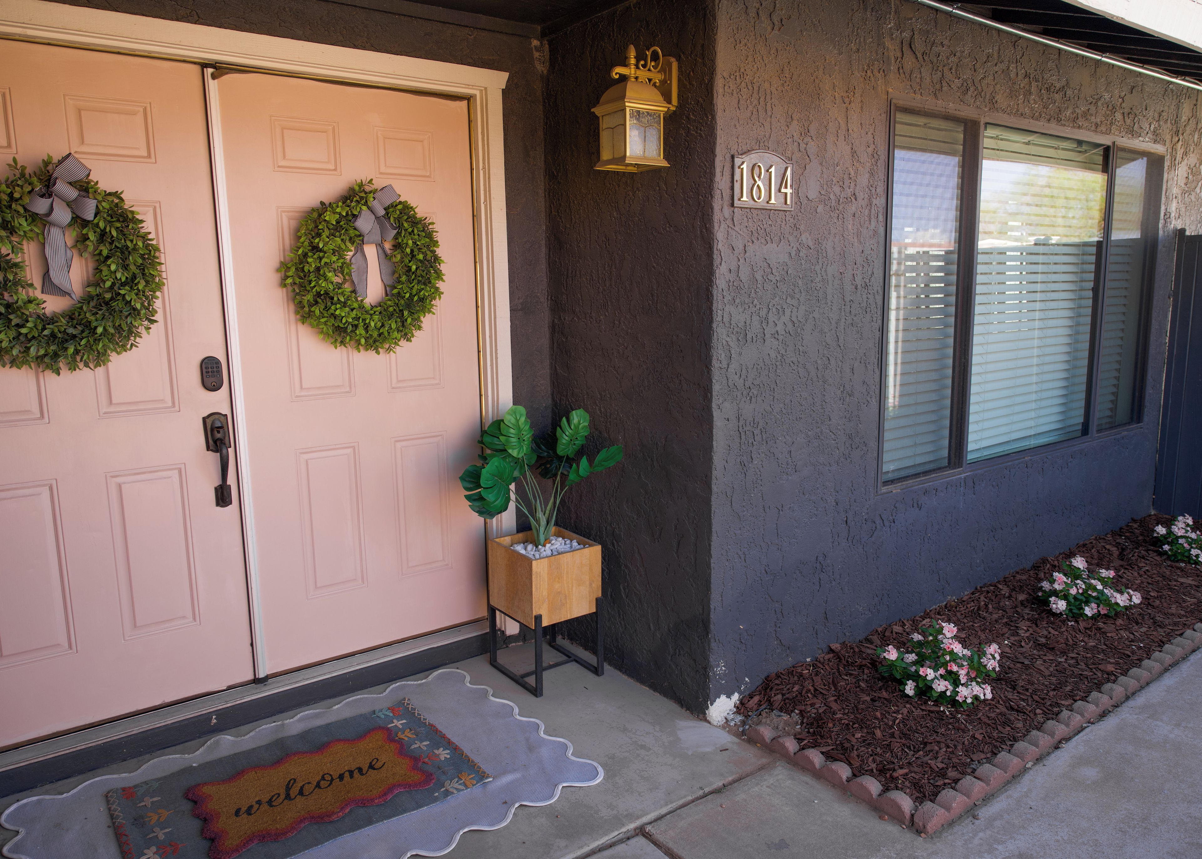 A house with a double door, a welcome mat, a number plaque, a potted plant, and a window with blinds.
