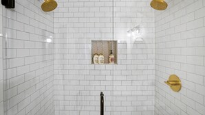 Un baño con paredes de azulejos blancos, una zona de ducha con detalles dorados y un estante con artículos de aseo.