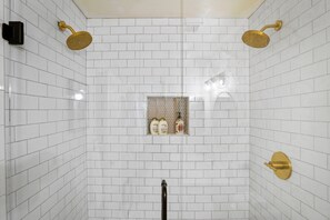 Un bagno con pareti rivestite di piastrelle bianche, zona doccia con accessori dorati e un piccolo ripiano con articoli da toilette.