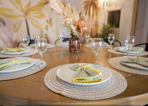 Una mesa de comedor dispuesta con platos, cubiertos y copas de vino, con manteles individuales y un jarrón de flores en el centro.
