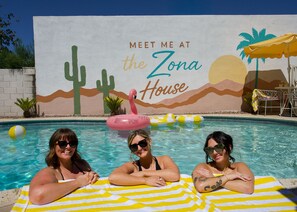 Un área de piscina con un mural que dice 'MEET ME AT the Zona House' e incluye un flotador con forma de flamenco.