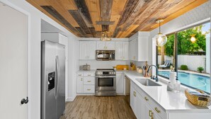 Una cocina con armarios blancos, electrodomésticos de acero inoxidable, techo de madera y vista a una piscina.